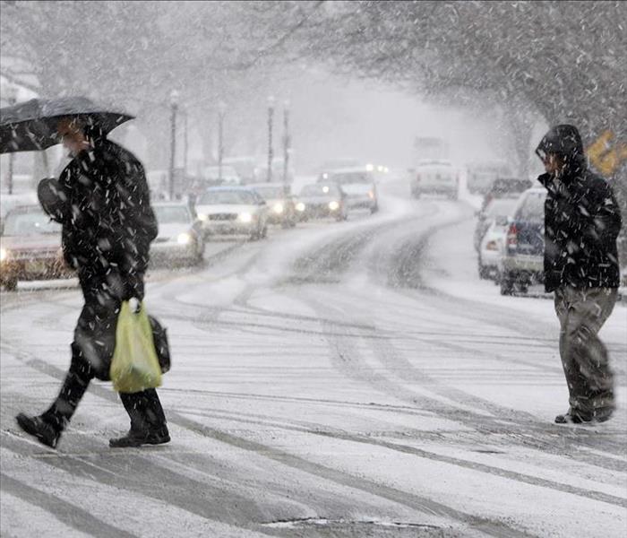 Two pedestrians crossing the street in the middle of a snowstorm.